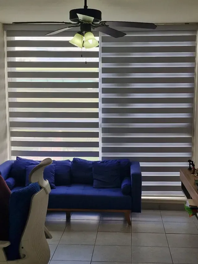 Sala de estar con sofá azul, ventilador de techo, suelo de baldosas y persianas de cebra que cubren una gran ventana.