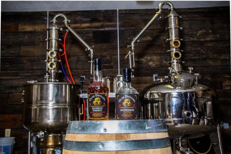 Two bottles of Old Southern Whiskey, bourbon and moonshine, sit on a wooden barrel in front of two metal stills.