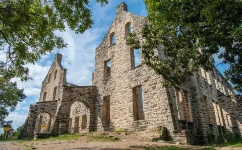 The stone ruins of a historic multi-story building stand under a blue sky, framed by trees.