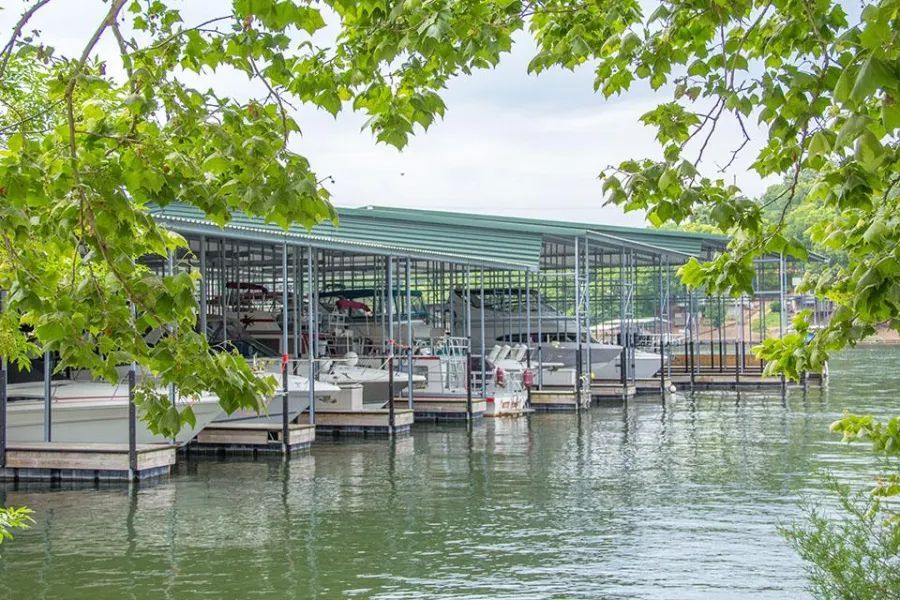 A metal covered boat dock with several boats moored under its roof on a calm lake, framed by green tree branches.