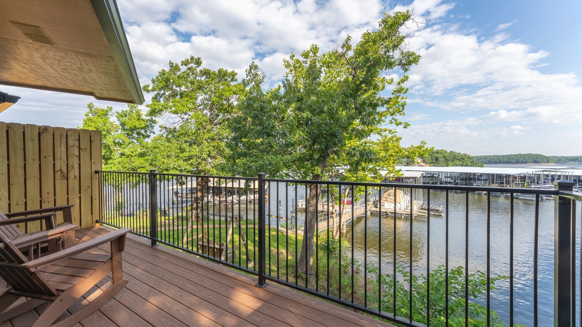 A wooden deck with chairs overlooking a lake with a marina, surrounded by trees under a partly cloudy sky.