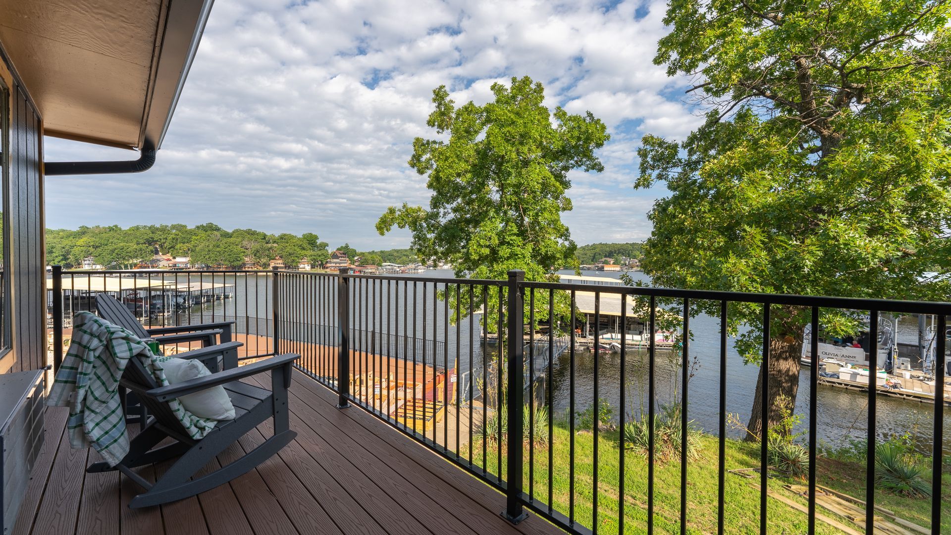 A wooden deck overlooking a lake with lush trees, a metal railing, and two rocking chairs with blankets.