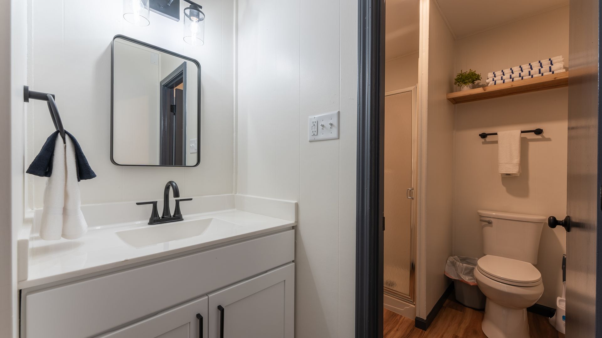 A modern bathroom with a white vanity, black fixtures, a rectangular mirror, and a toilet in an adjacent nook.