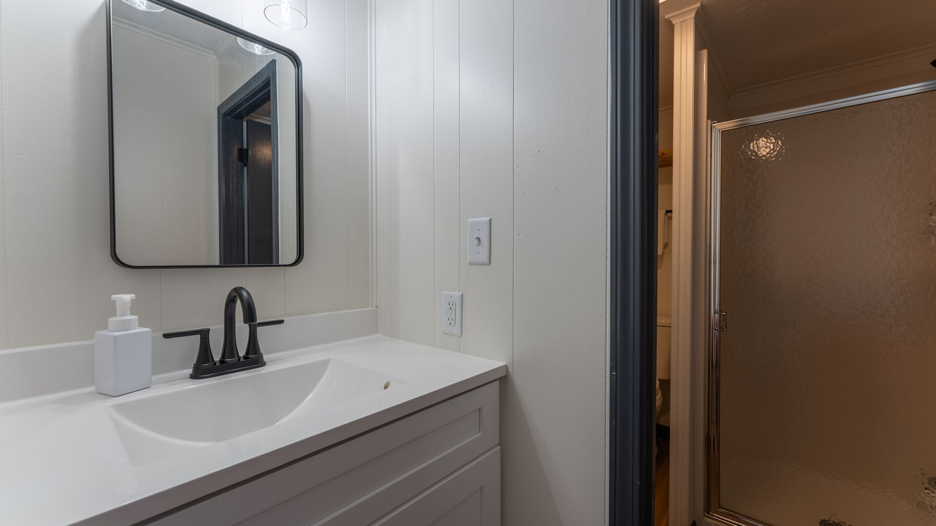 A white bathroom vanity with a modern black faucet and rectangular mirror next to a walk-in shower.