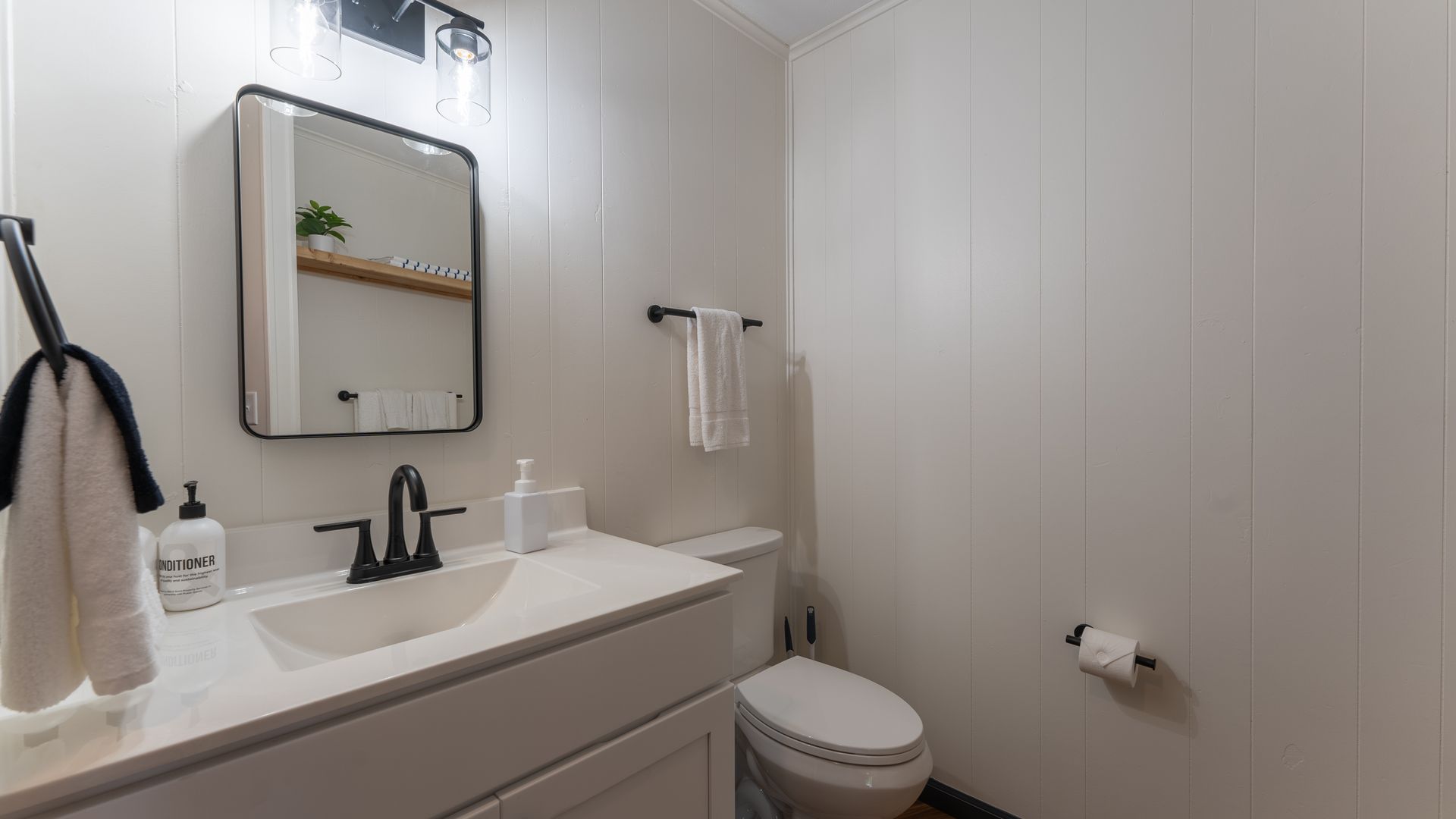 A modern bathroom featuring a white vanity with a black faucet, rectangular mirror, and toilet against light paneled walls.