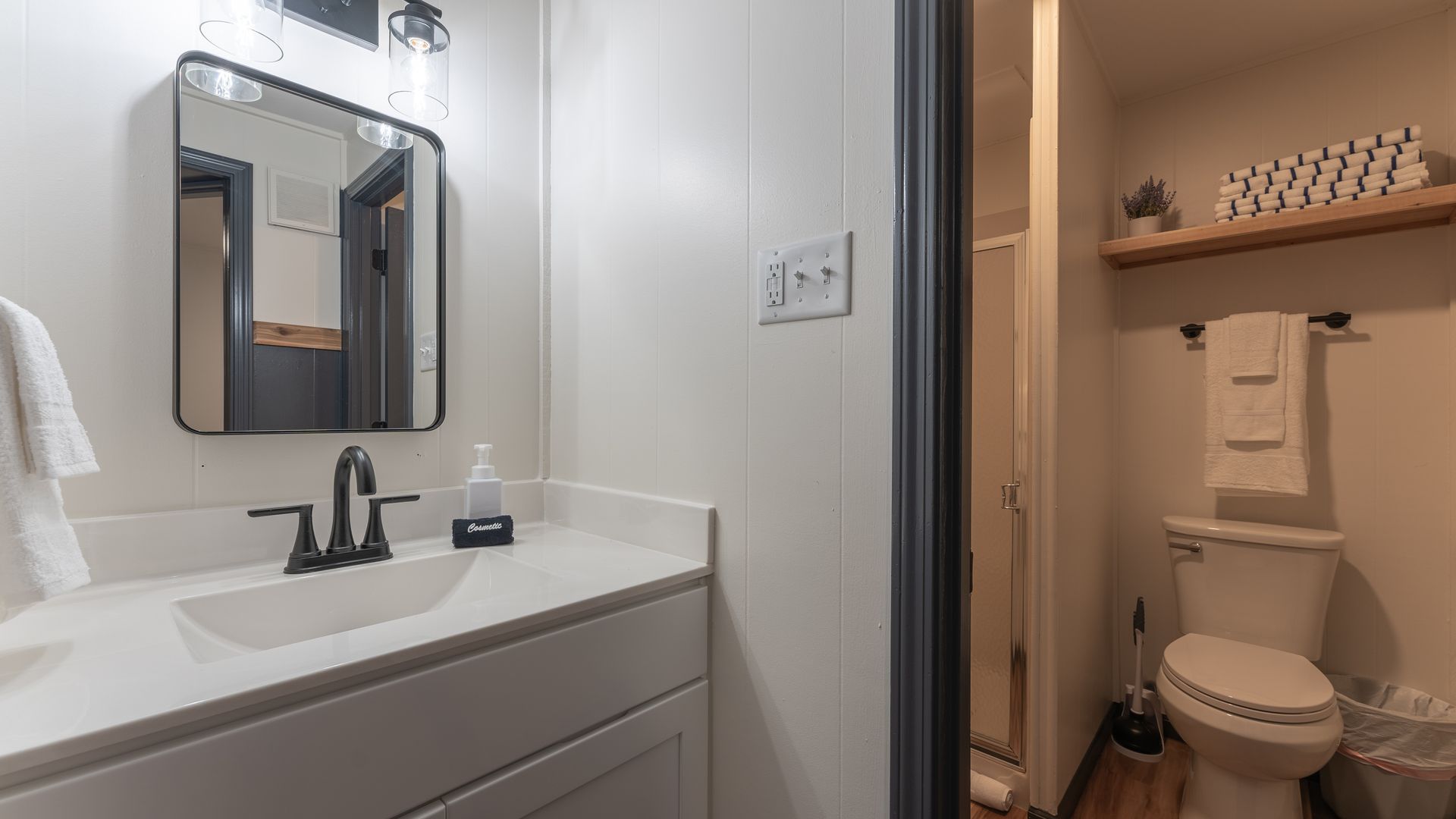 A modern bathroom featuring a white vanity with a dark faucet, a rectangular mirror, a toilet, and a wooden shelf above.