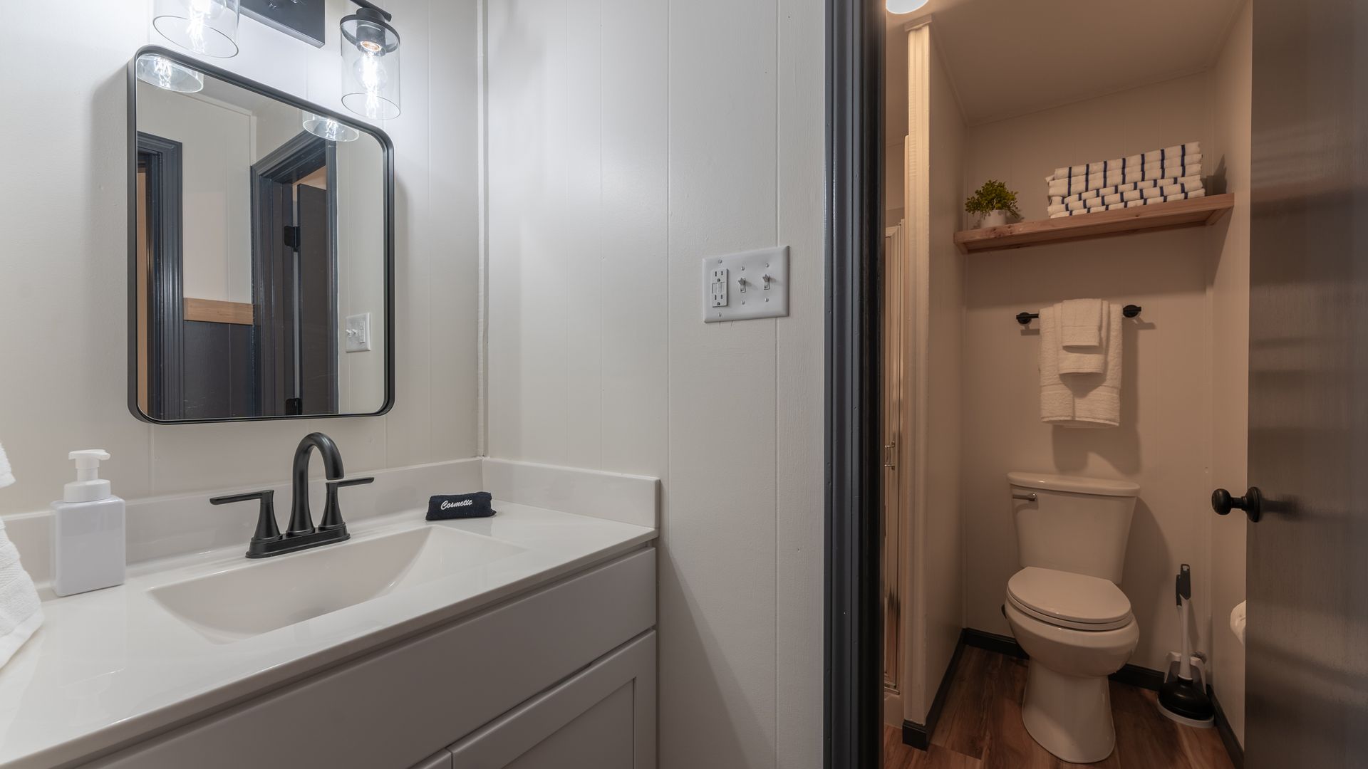 A modern bathroom features a vanity with a white countertop and dark faucet, opening into a small room with a toilet.