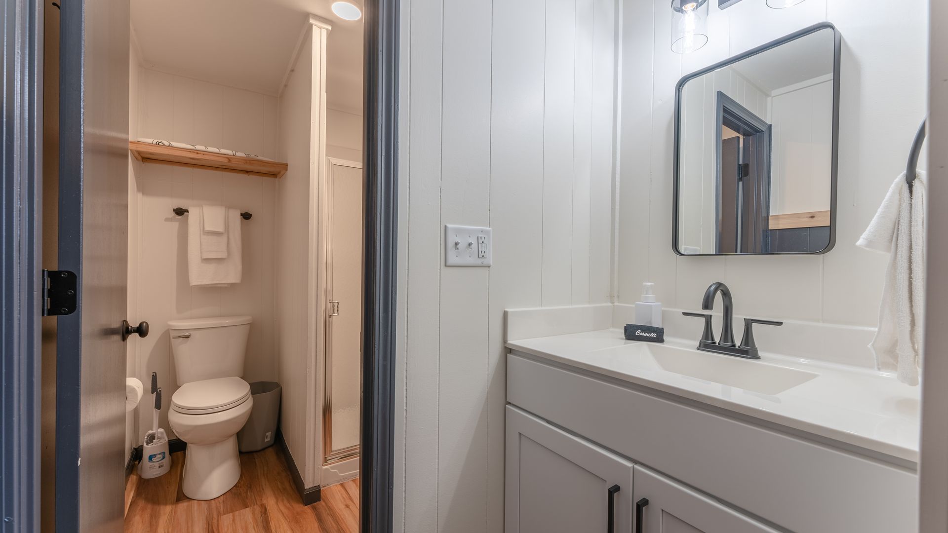 A bright, white bathroom with a vanity, mirror, and a separate room containing a toilet and shower with wood-style floors.