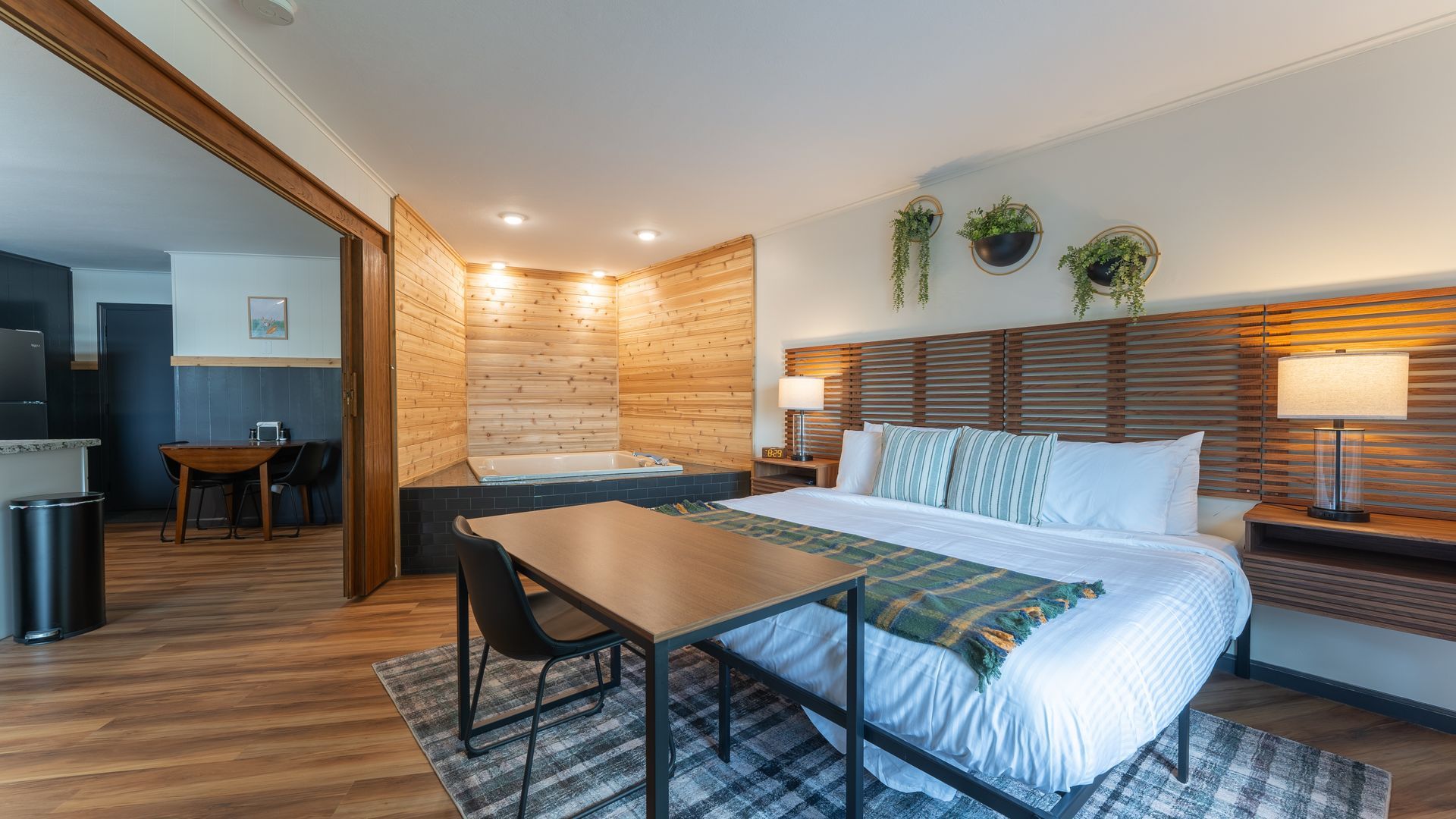 A hotel room with a large bed, wooden headboard, small table, and a partitioned kitchenette area with a dining set.