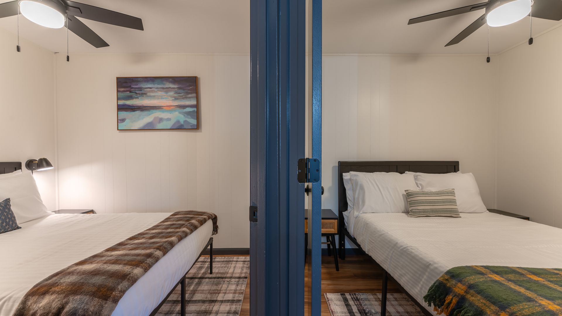 Two bedrooms separated by a blue door frame, each featuring a bed with white linens and a plaid runner.