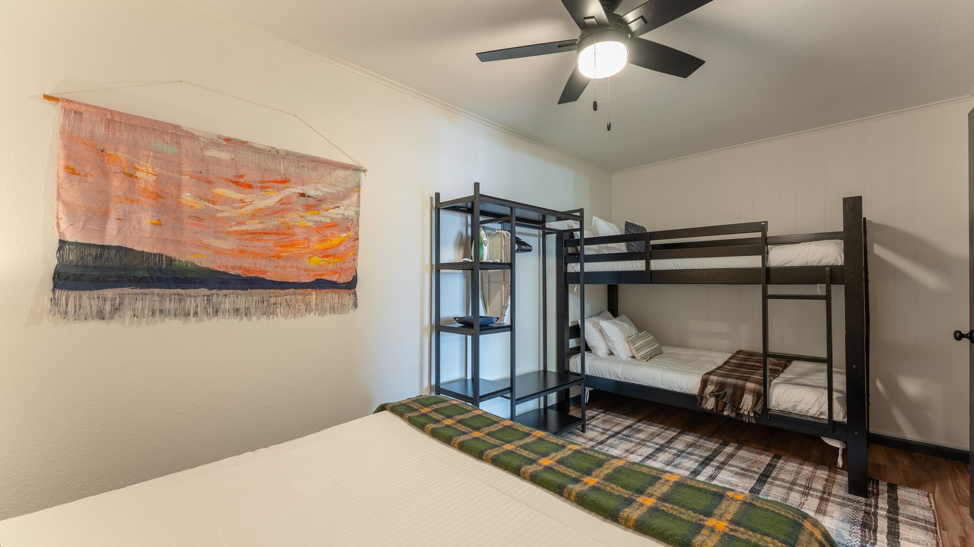 A bedroom features a queen bed, a black metal bunk bed, a matching shelving unit, and a sunset wall tapestry.