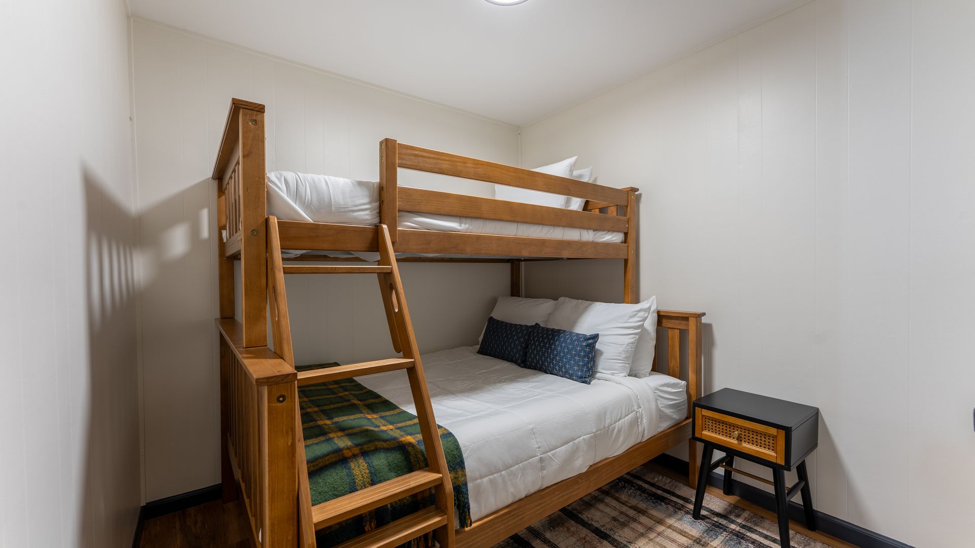 A wooden bunk bed with a top twin and bottom full mattress, white bedding, and a small dark bedside table in a room.