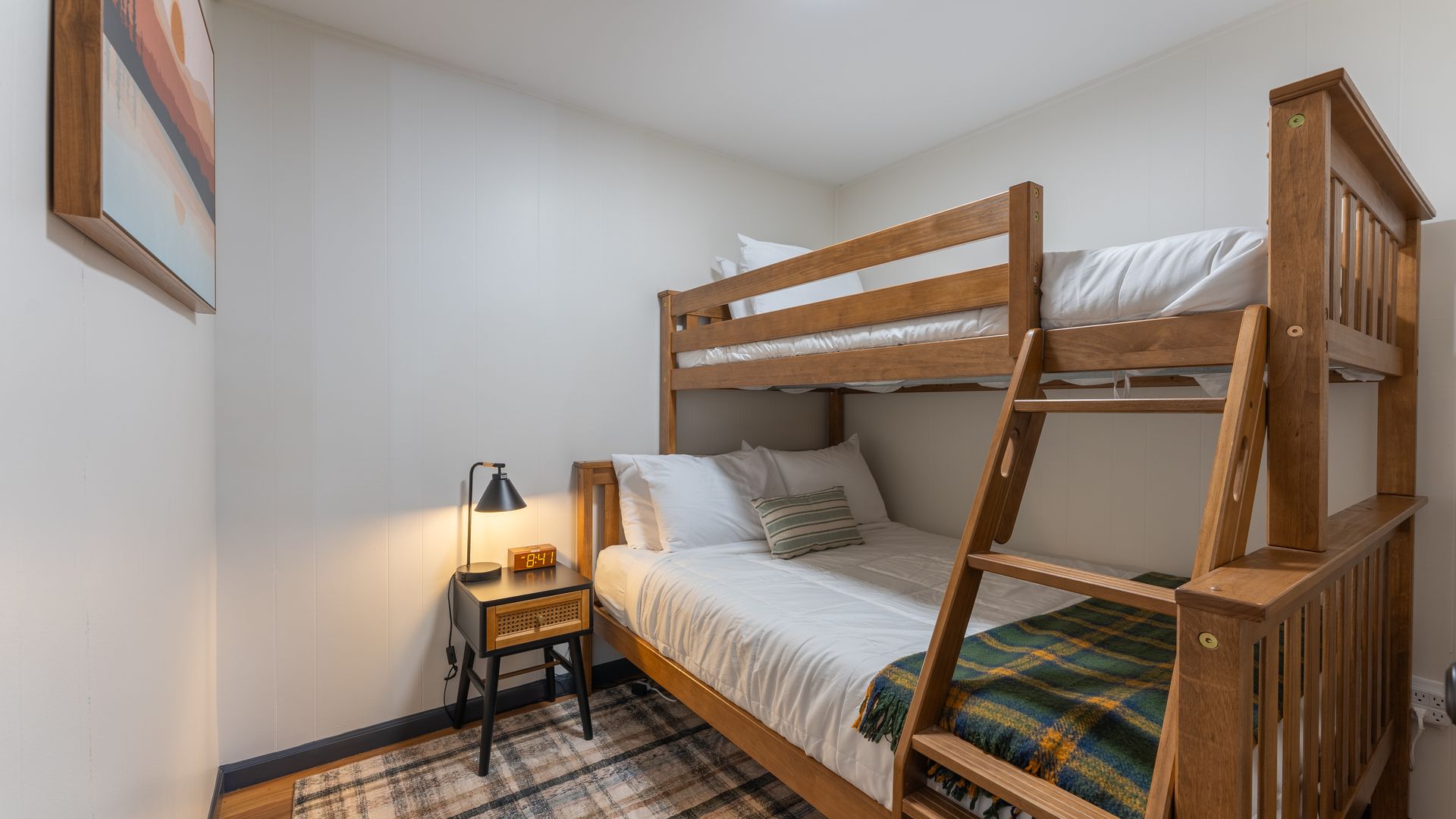 A wooden bunk bed with white bedding, a plaid throw, and a small bedside table with a lamp in a simple, neutral room.