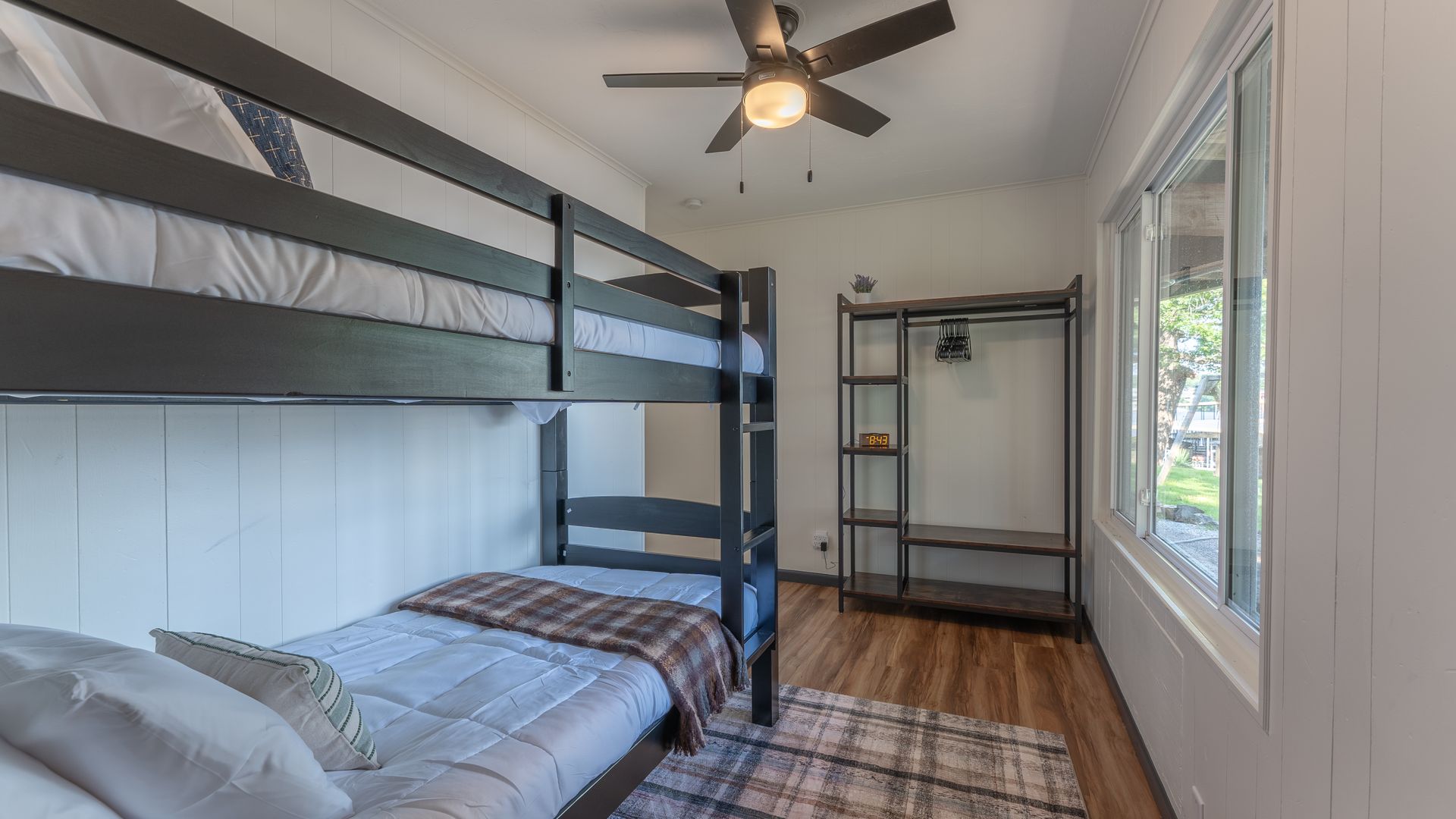 A bedroom with a dark wooden bunk bed, an open metal shelving unit, and a window, set on wood-look flooring.
