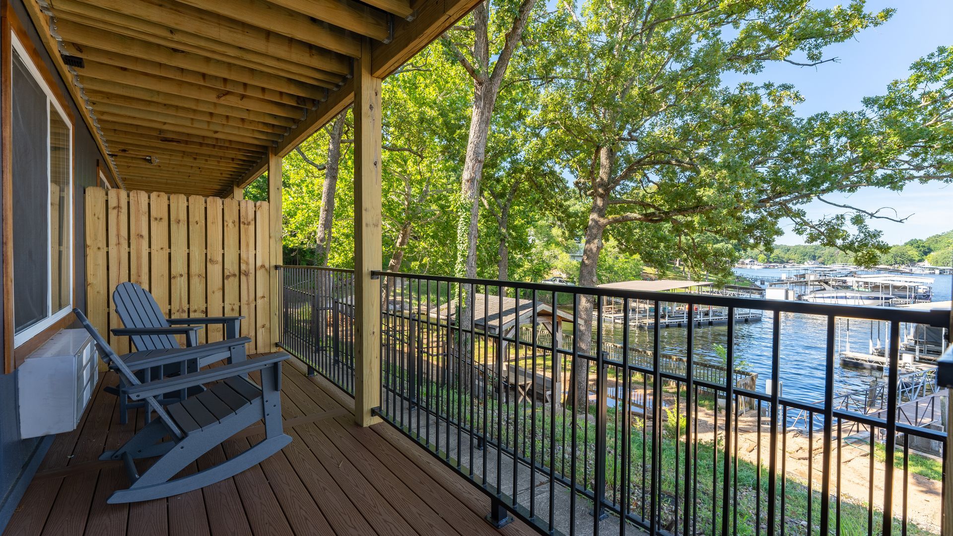 A covered wooden deck with two rocking chairs, overlooking a lake with boat docks surrounded by green trees.
