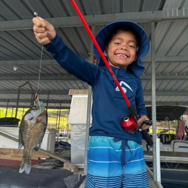 Smiling child in a blue hooded jacket holds a fishing rod with a caught fish hanging beside them.