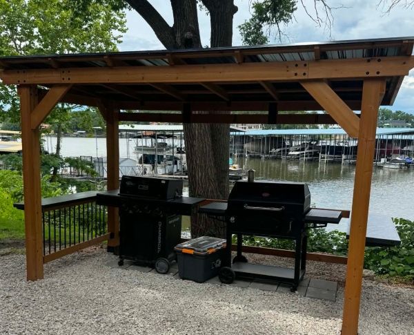 Covered outdoor grill station beside a lake, with black grills under a wooden shelter and docked boats in the background