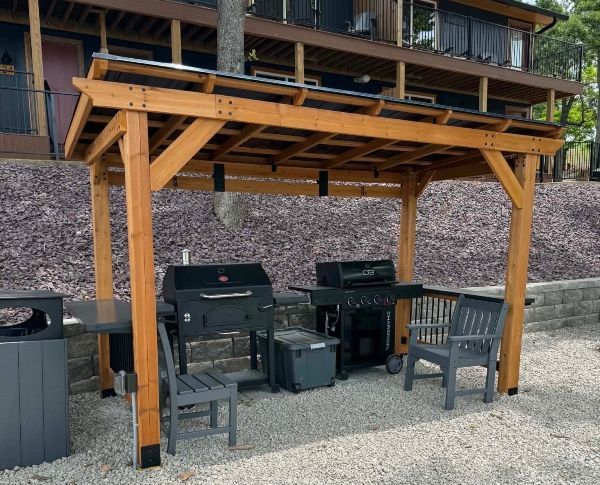 Outdoor covered grilling area with two black grills and several patio chairs on gravel.