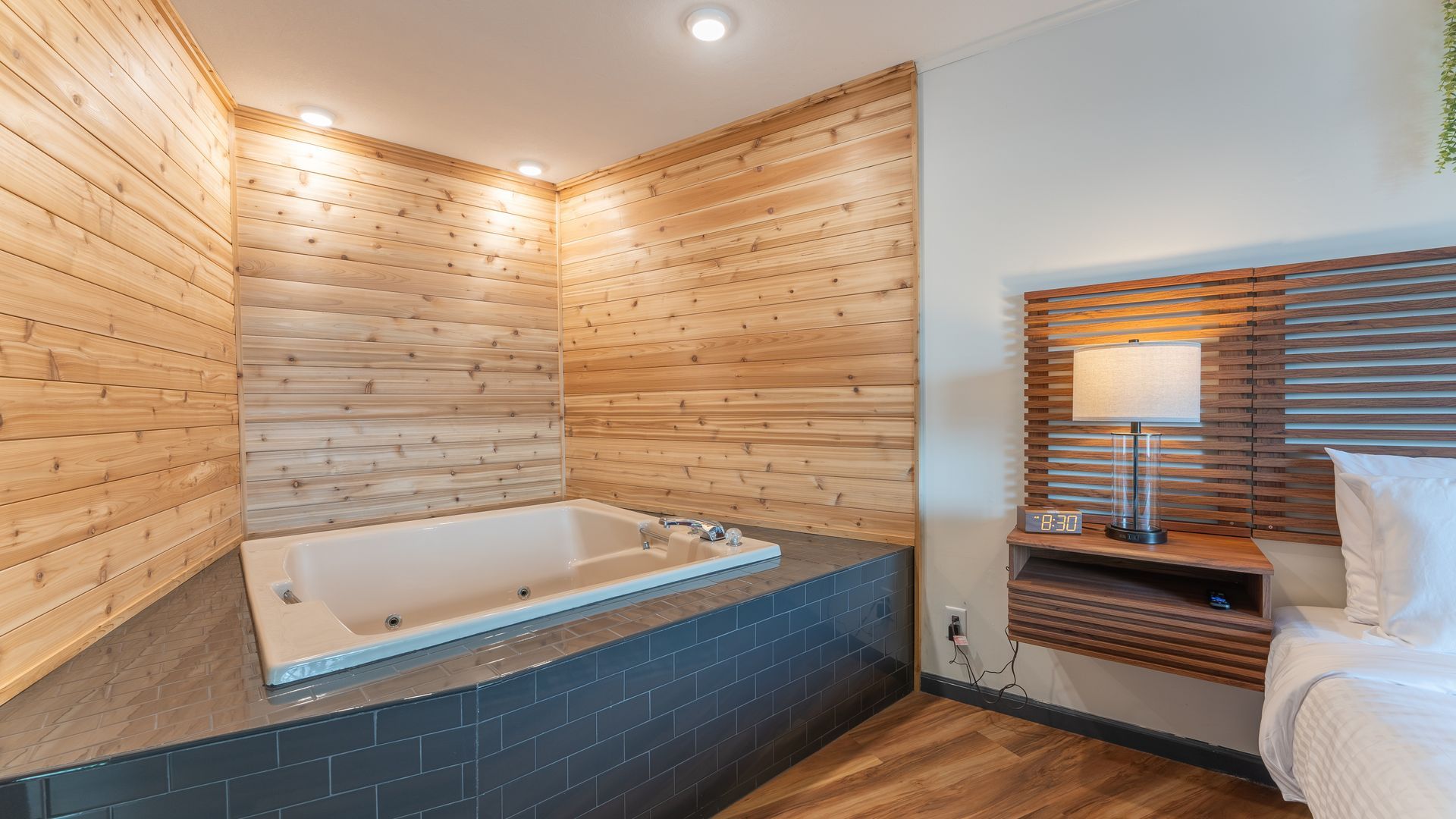A corner whirlpool bathtub with wood-paneled walls and dark tiles next to a bedside lamp and headboard in a hotel room.