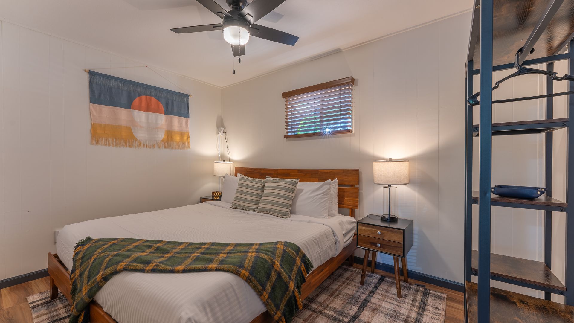 A bedroom with a queen bed featuring a green plaid throw, wooden headboard, two nightstands, and a hanging wall tapestry.