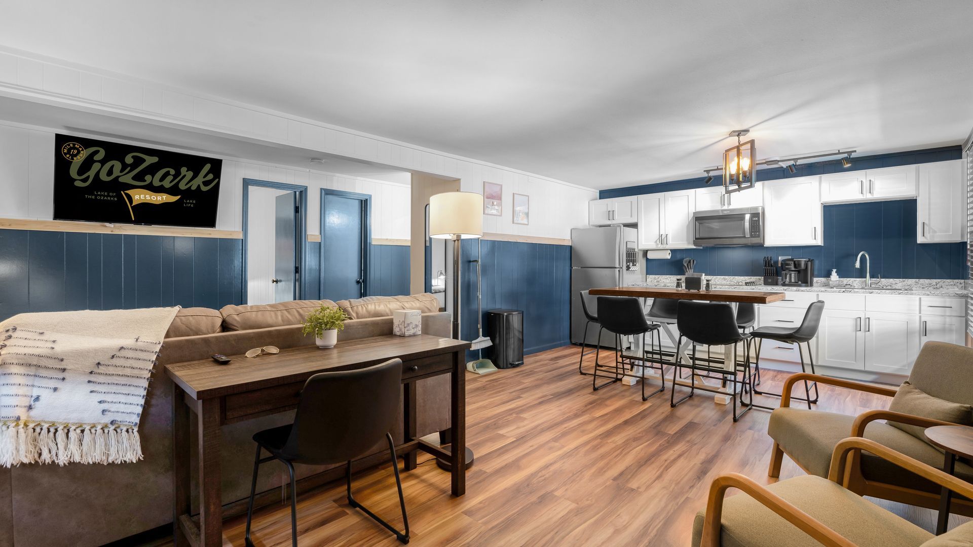A modern living area with a kitchenette, brown desk, sofa, and chairs on wood flooring, featuring navy blue wainscoting.