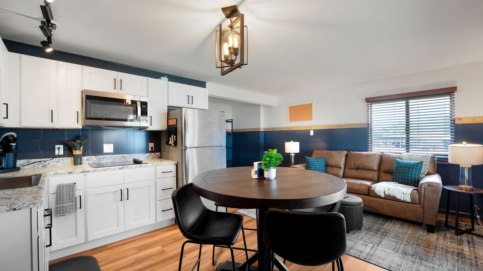 Open-concept kitchen and living area featuring white cabinets, a dark blue backsplash, dining table, and brown sofa.
