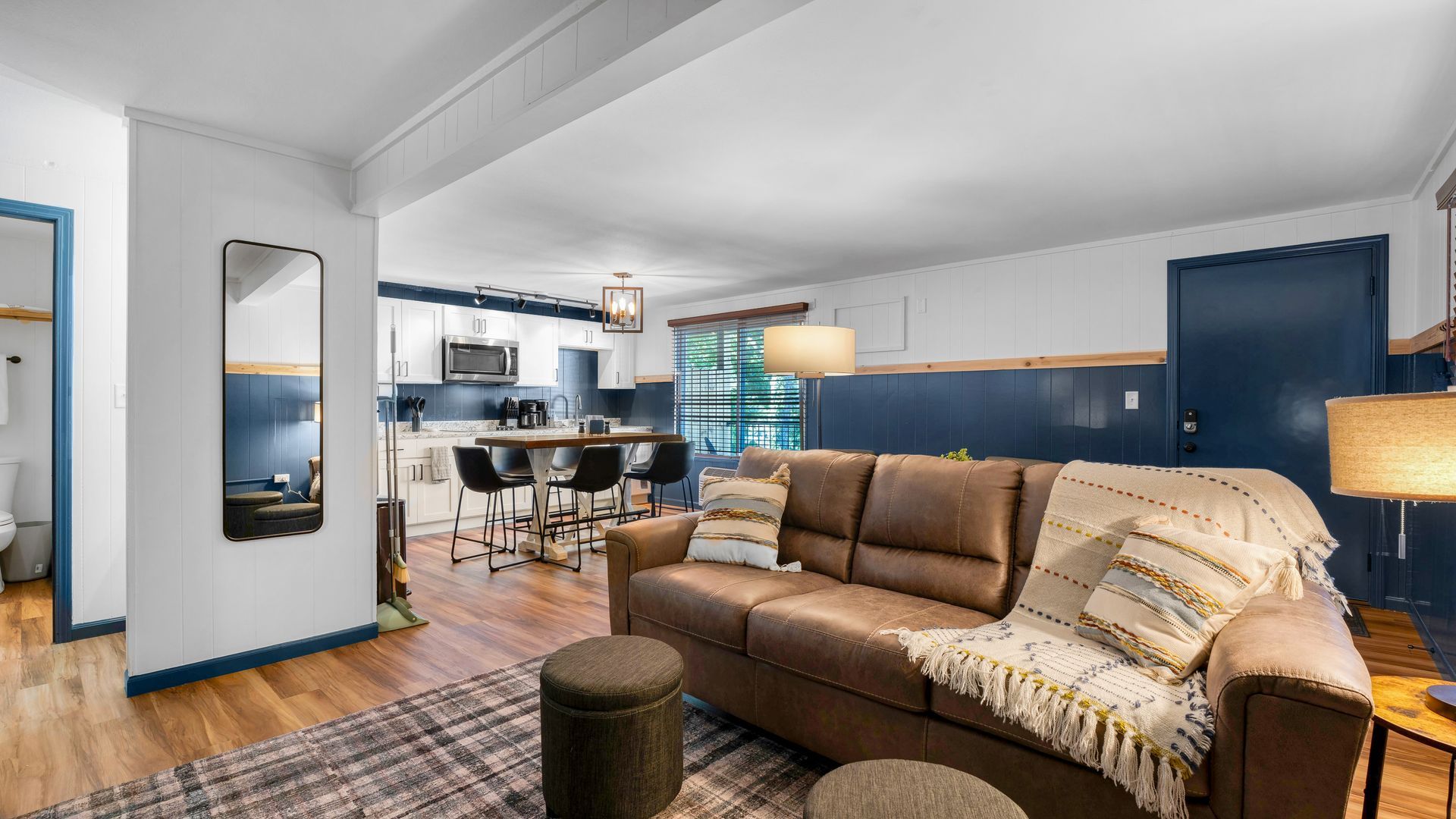 A brown leather sofa sits on a plaid rug in a living room with dark blue wainscoting, leading to a white kitchen.