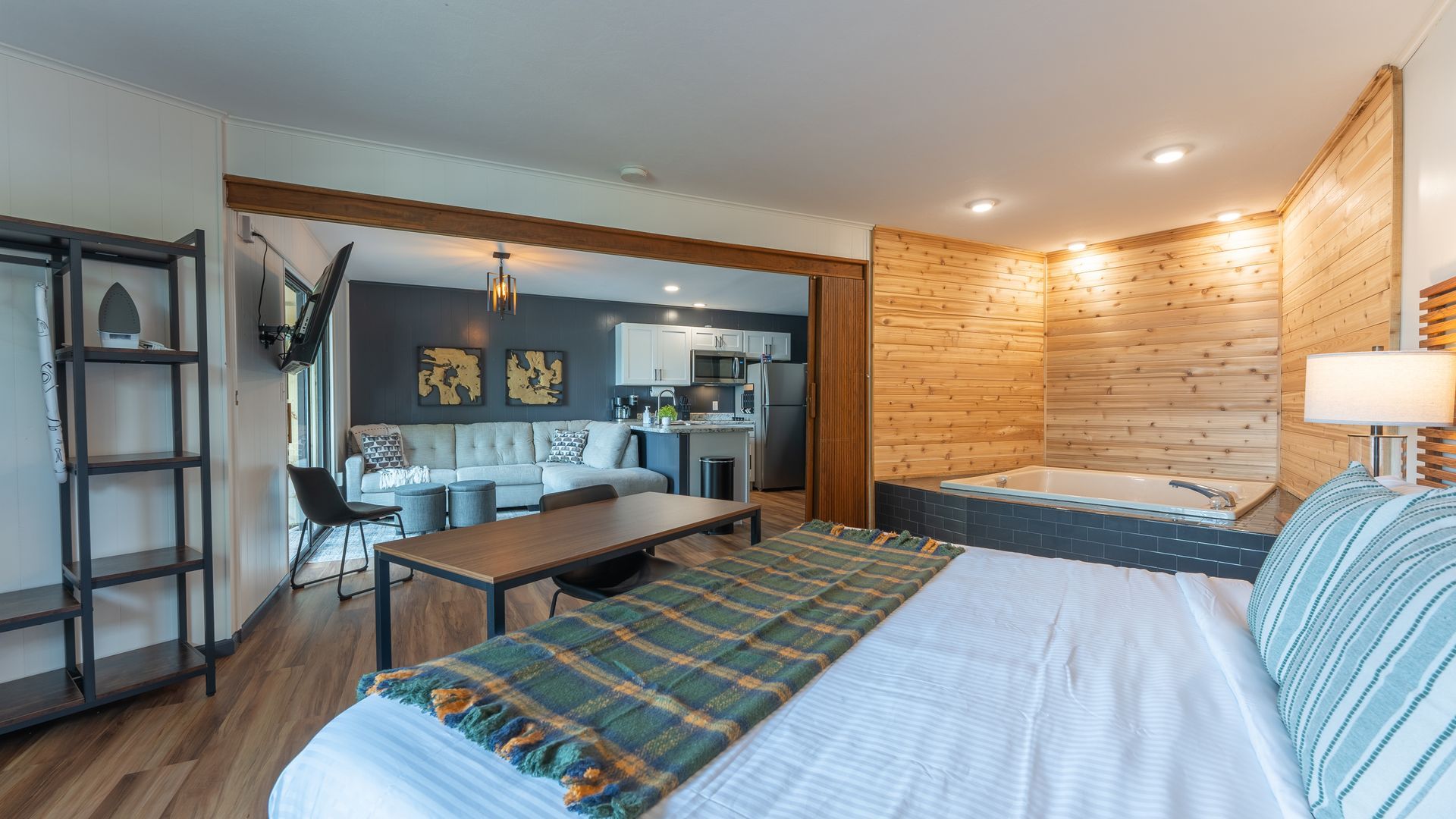 A hotel room featuring a bed with a plaid throw, a wooden jetted tub, and a view into a kitchen and living area.