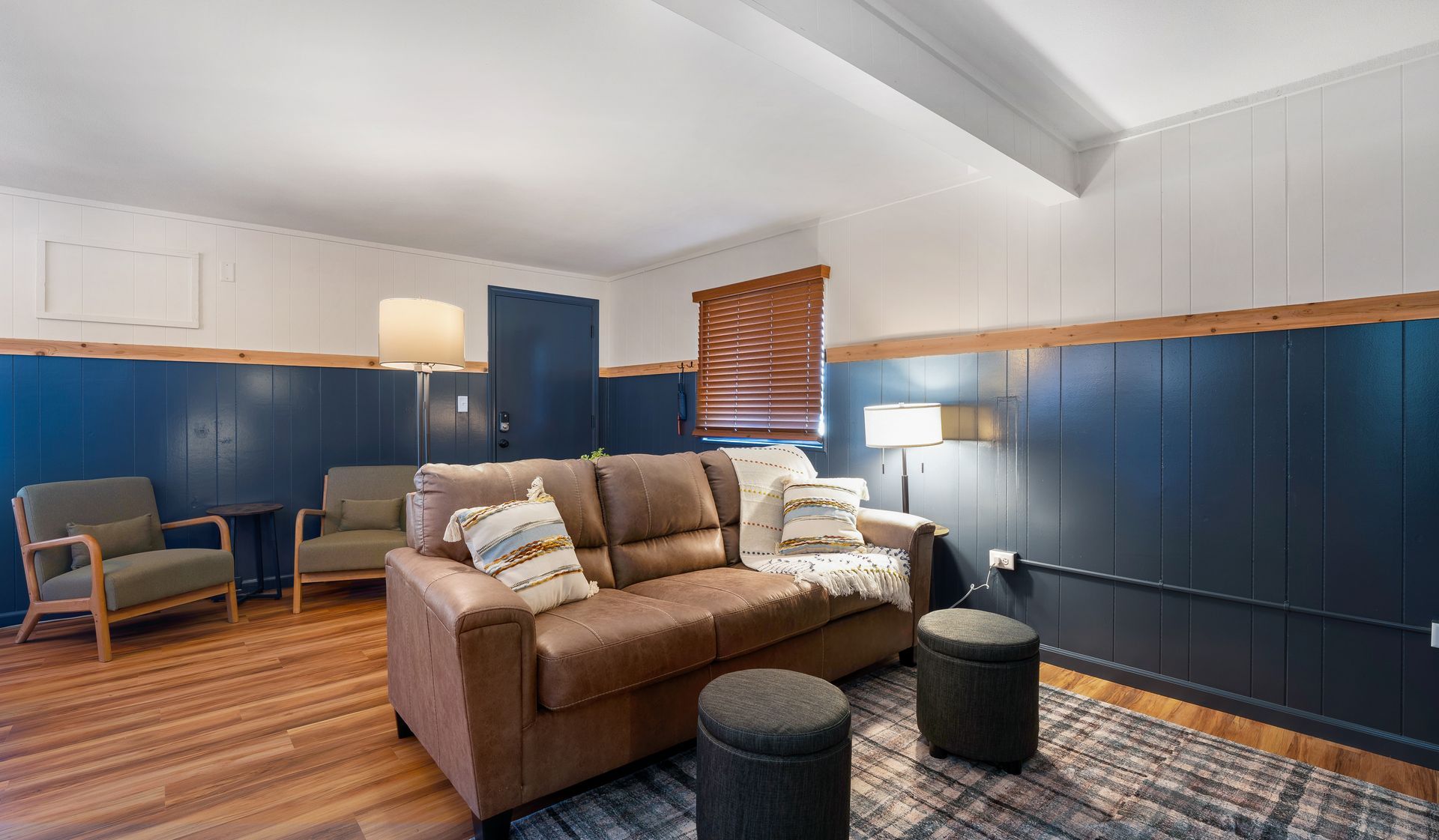 A cozy living room with a brown leather sofa, two grey chairs, dark blue paneled walls, and wooden floors.