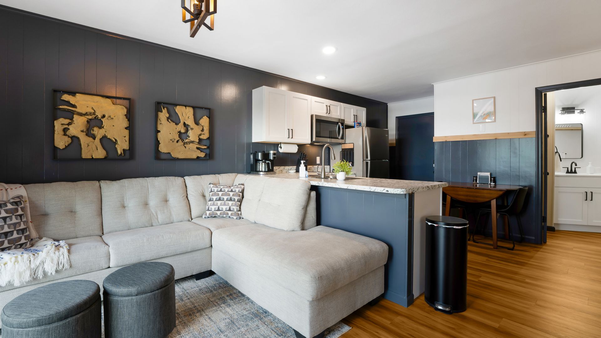 A light-colored sectional sofa faces a dark accent wall with two gold art pieces in a modern kitchen and living space.