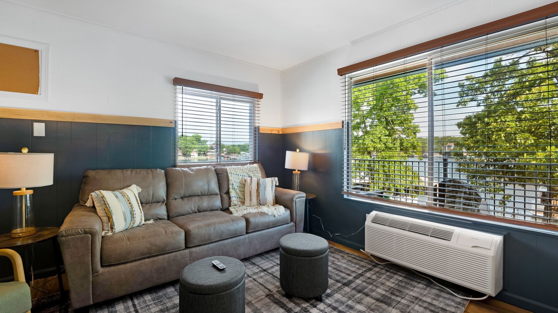A living room with a brown sofa, two ottomans, and side tables against blue walls with white upper trim and large windows.