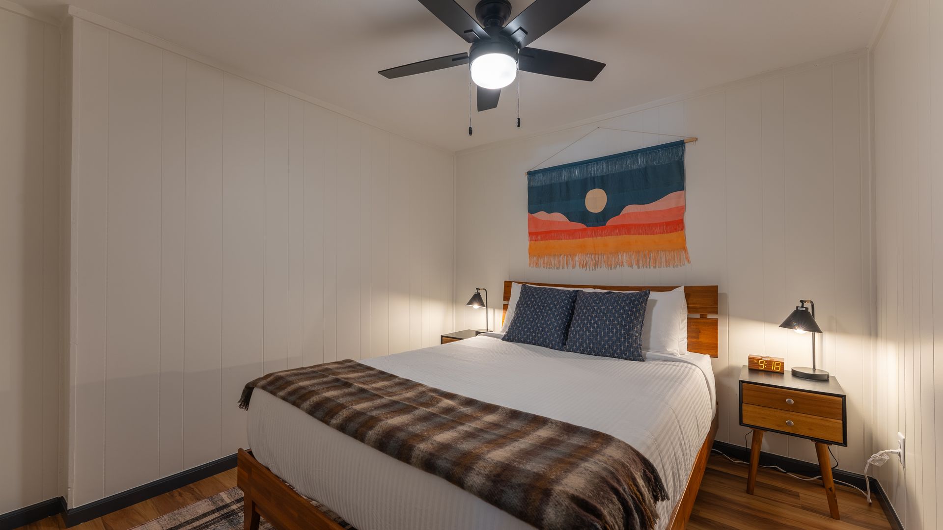 A minimalist bedroom featuring a bed with a plaid throw, two nightstands with lamps, and a landscape tapestry on the wall.