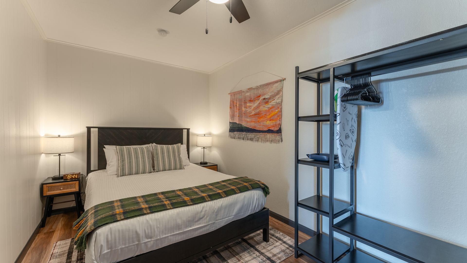 A bedroom featuring a bed with a plaid runner, two bedside lamps, a wall tapestry, and an open metal clothing rack.