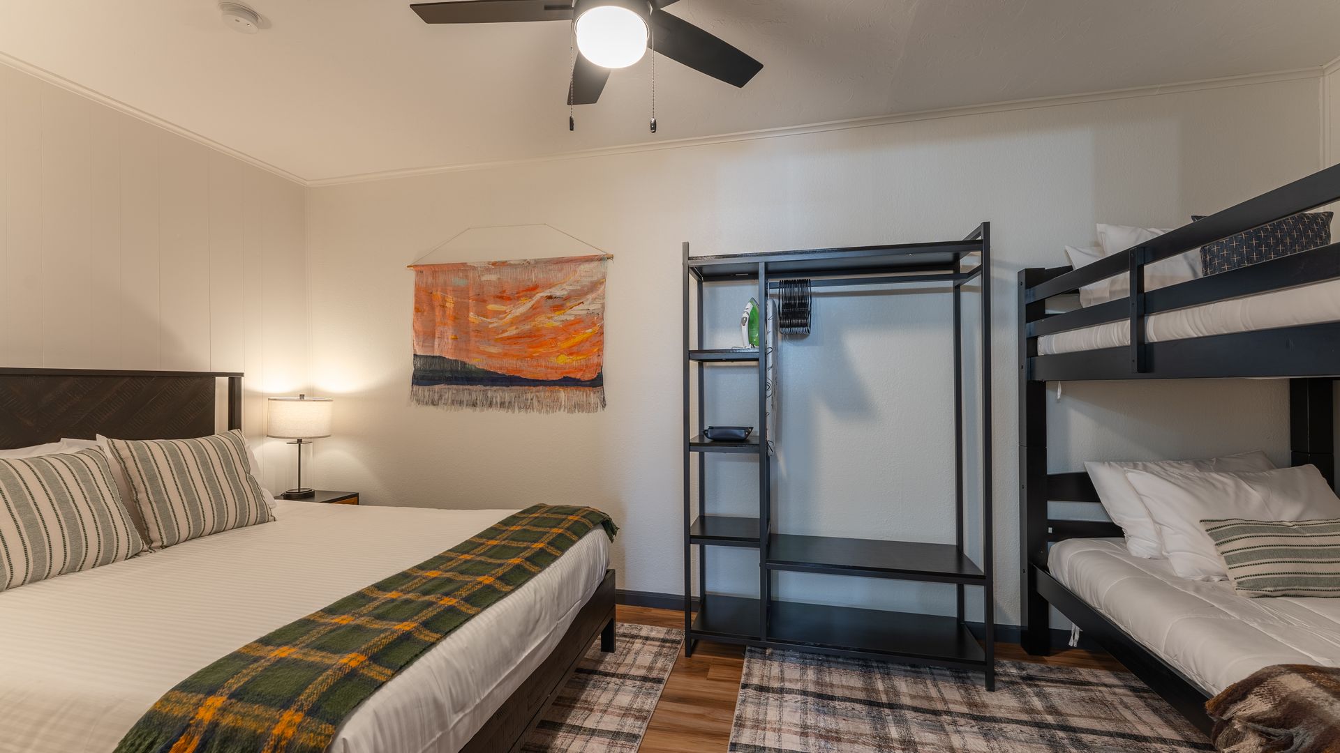 A bedroom with a queen bed, a black metal open wardrobe, and a bunk bed, featuring wood floors and neutral decor.