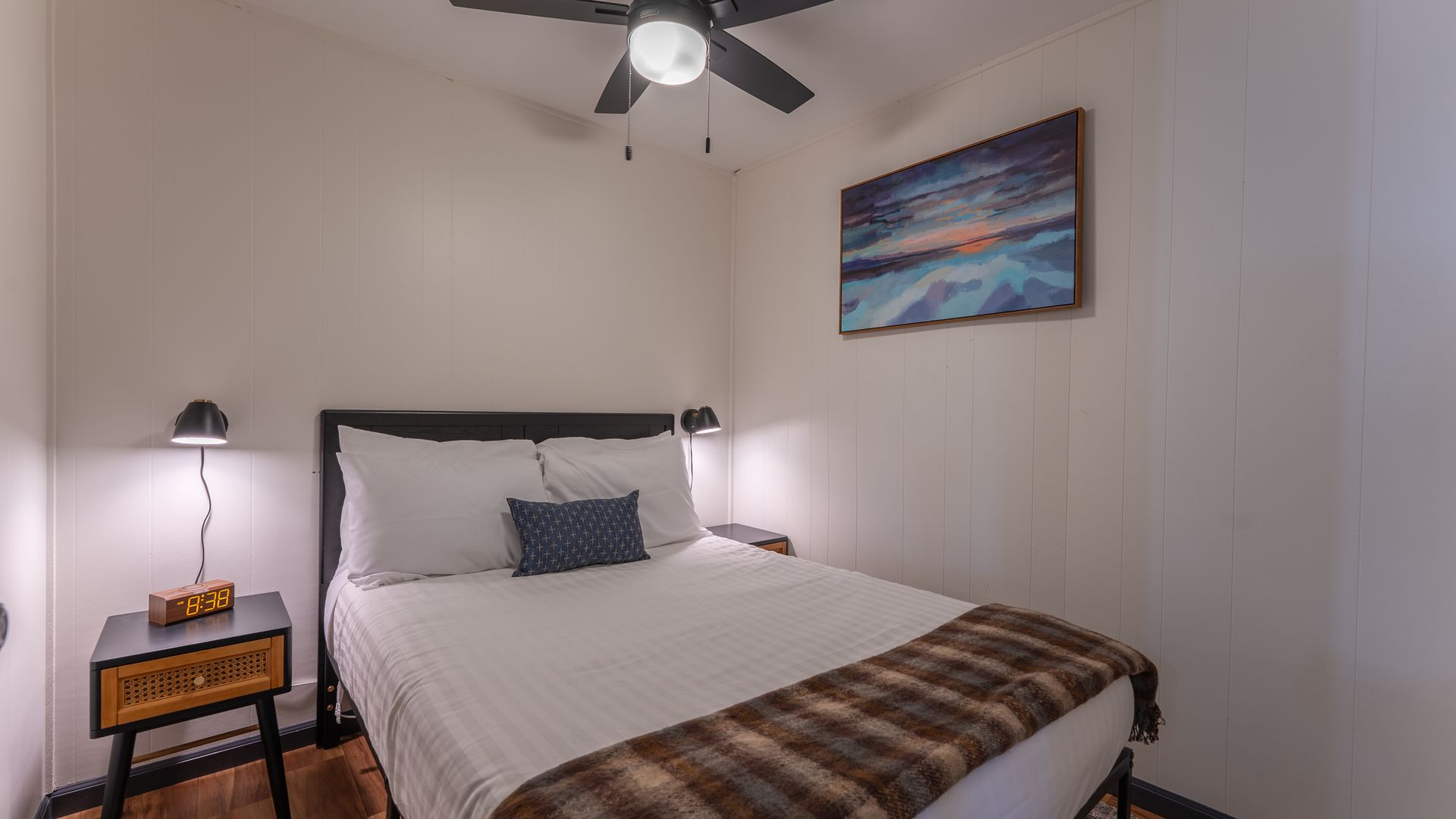 A cozy bedroom featuring a queen bed with white linens and a plaid throw blanket, a small bedside table, and wall art.