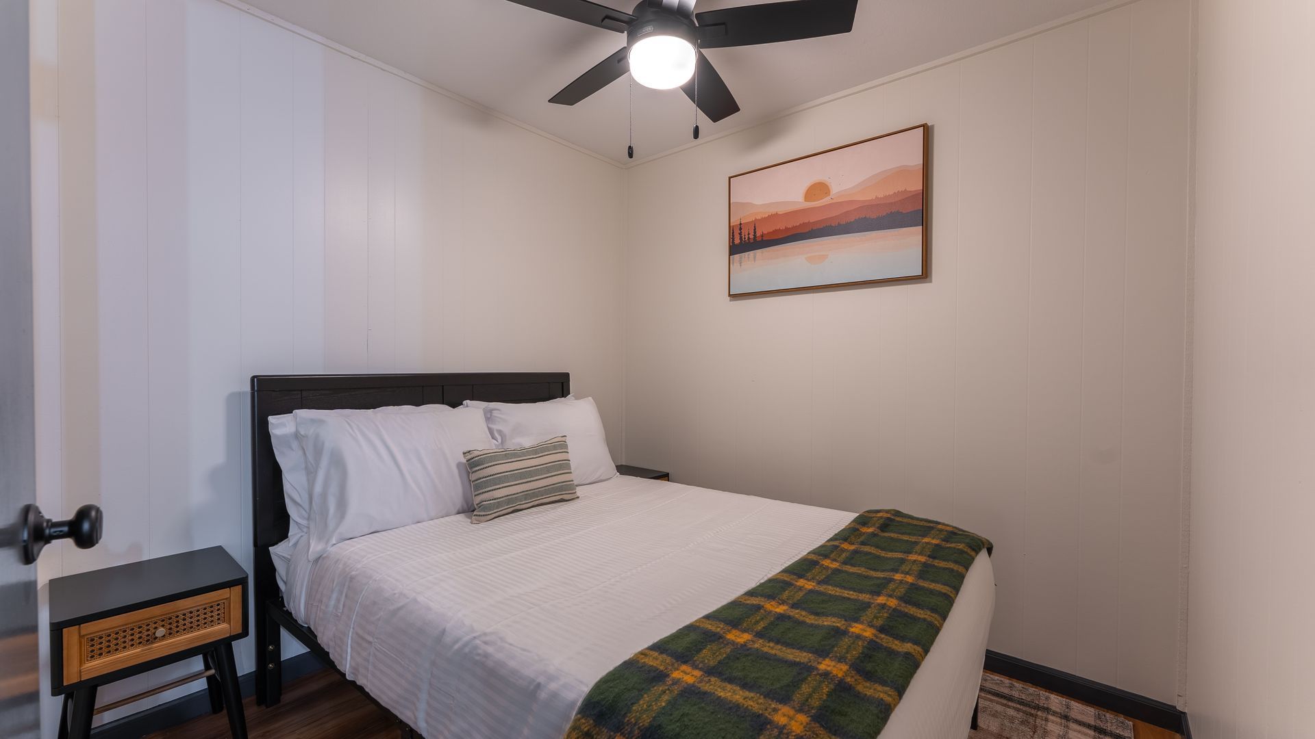 A simple bedroom with a bed featuring white bedding and a green plaid throw, a ceiling fan, and landscape wall art.