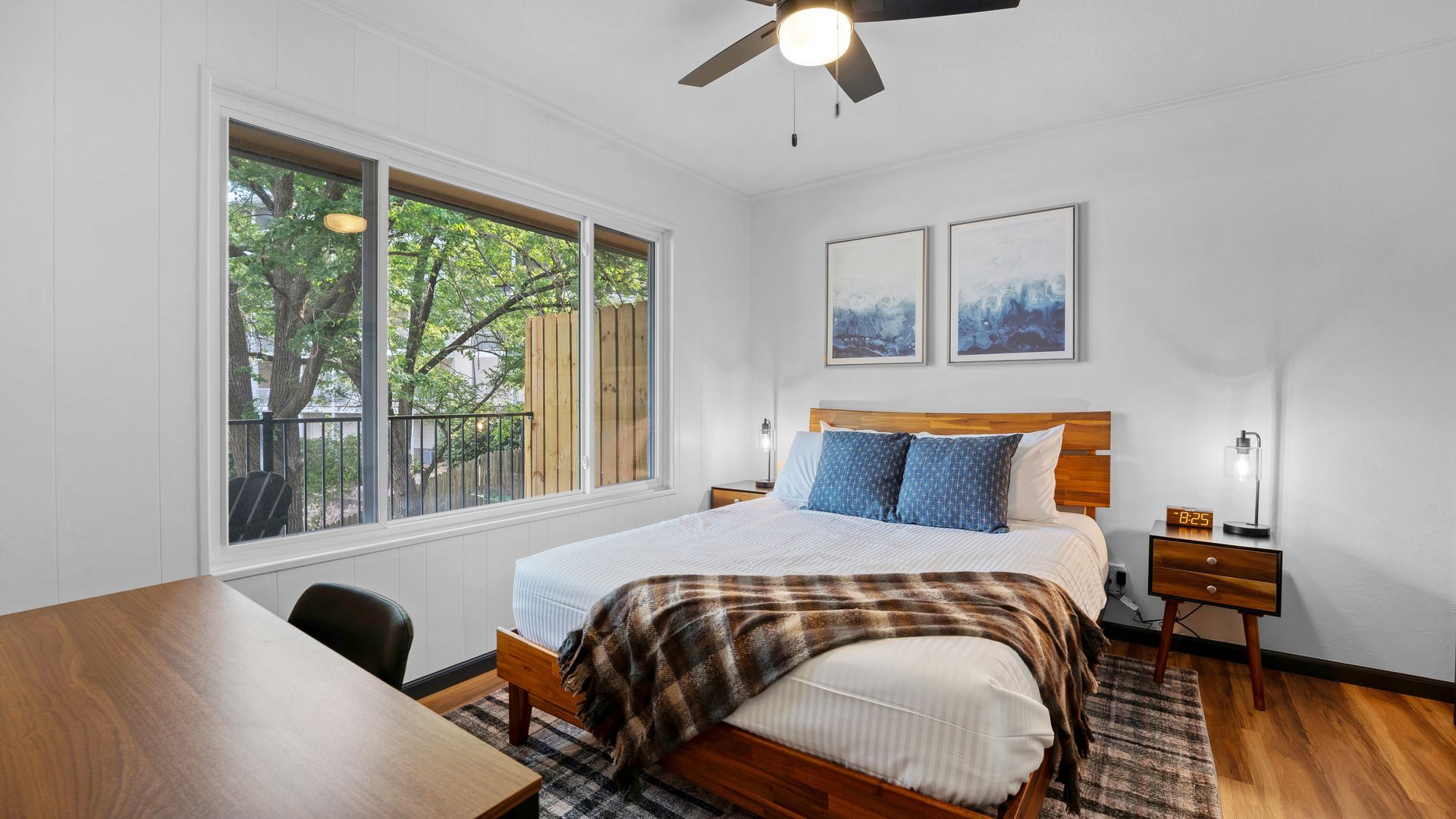 A bedroom with a wooden bed, brown plaid blanket, and blue pillows near a large window overlooking trees.