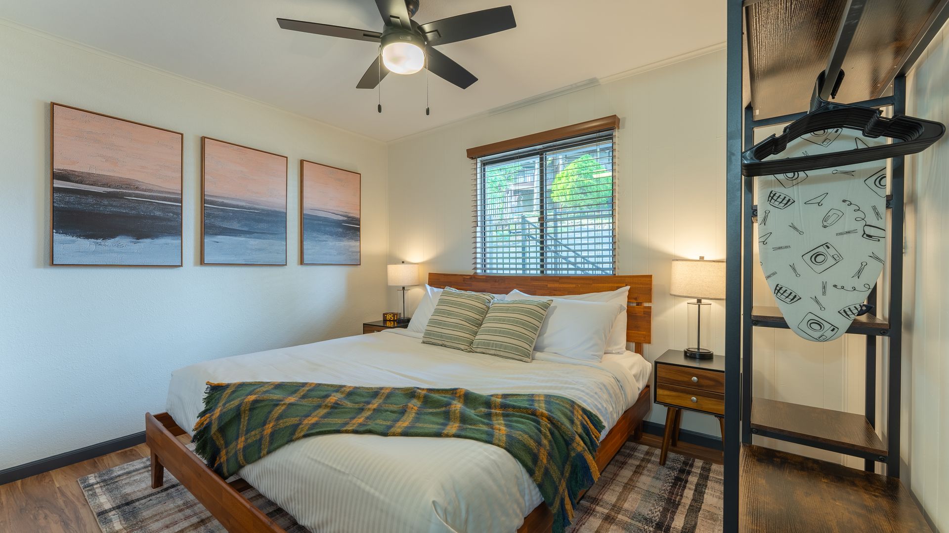 A bedroom with a bed featuring a green plaid blanket, three landscape paintings on the wall, and an open metal closet.