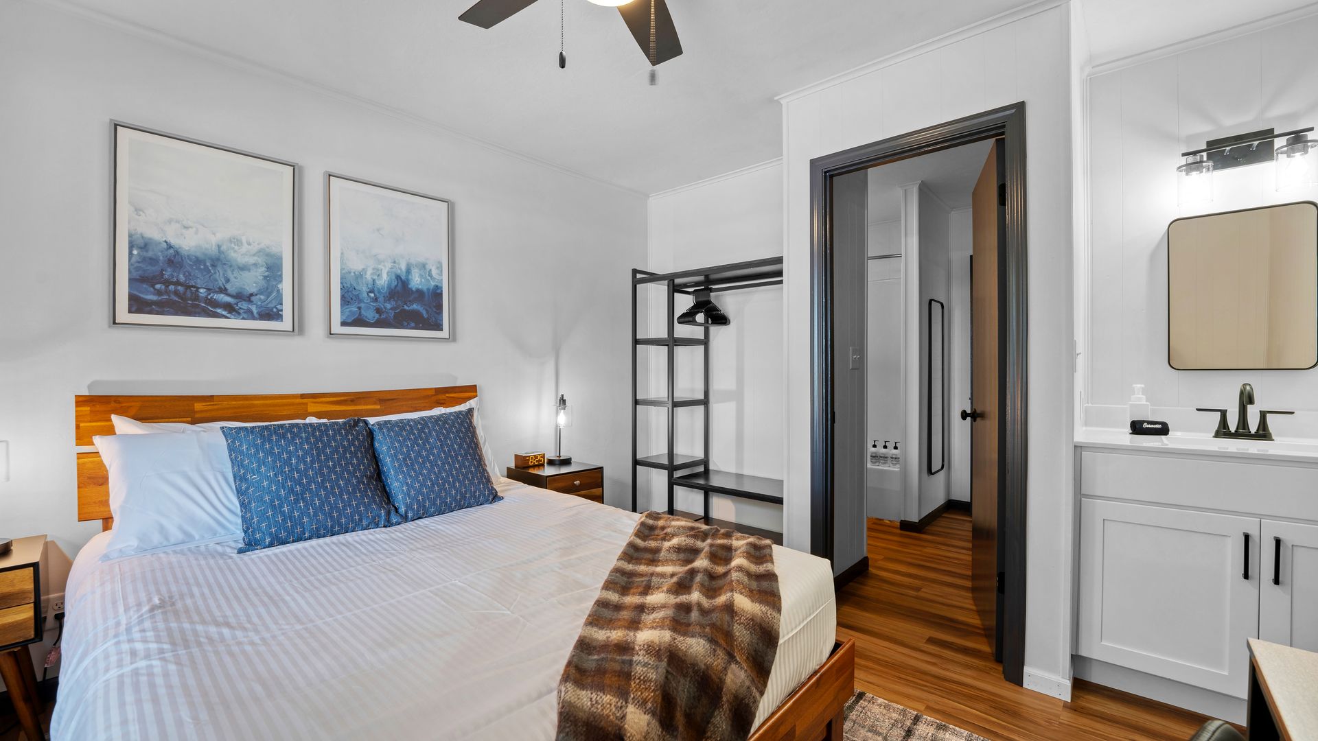 A bedroom with a queen bed, blue decorative pillows, a clothing rack, an open doorway, and an adjacent bathroom vanity.