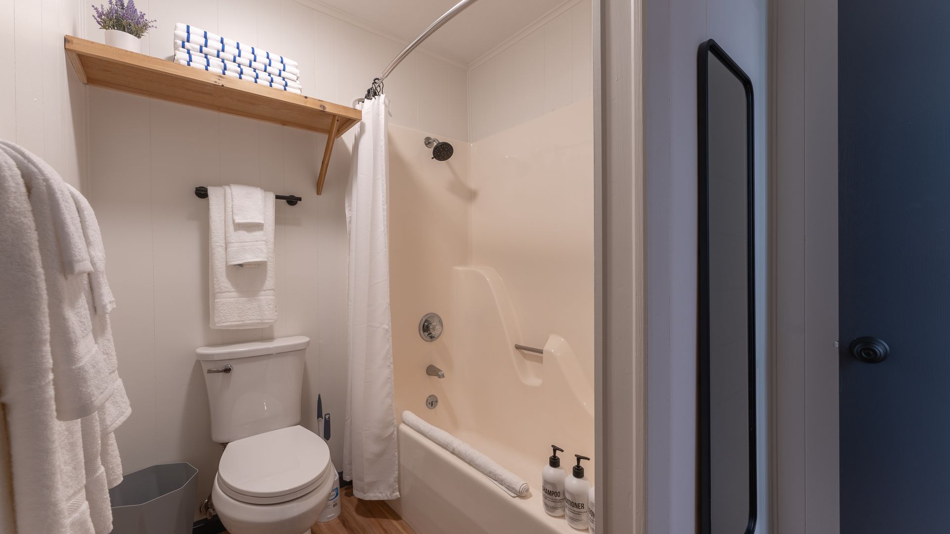 A small bathroom featuring a toilet, a shower-tub combo with a white curtain, and an open wooden shelf above the toilet.