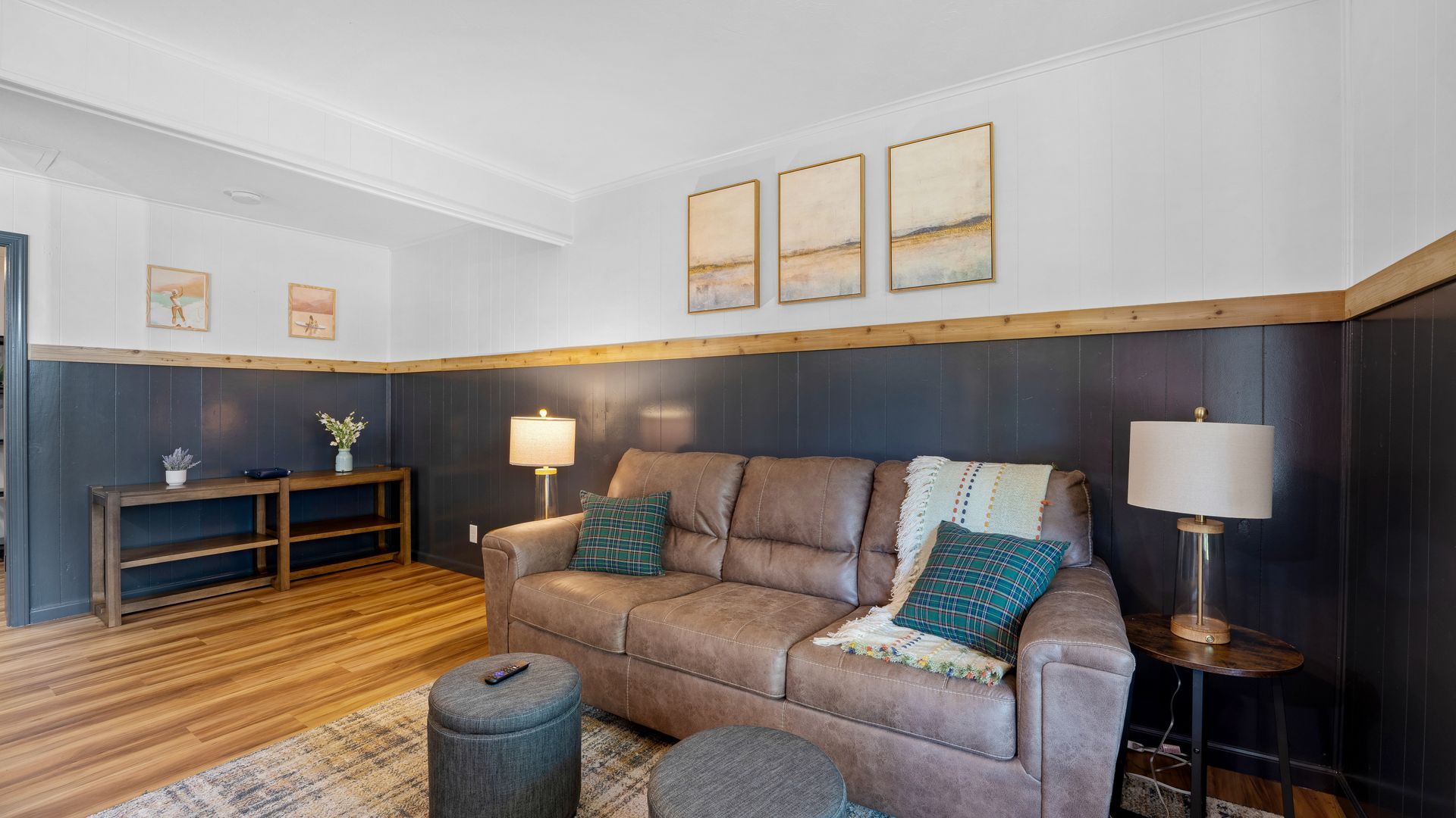 A living room with a brown sofa, gray ottomans, a wood table, dark paneled walls, and three framed prints above the sofa.