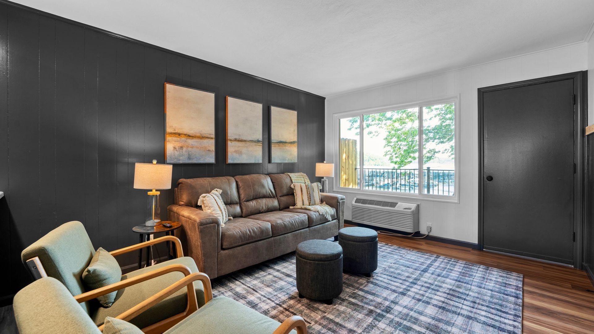 A modern living room with a gray wall, brown sofa, two light green armchairs, and a plaid rug on a wood floor.