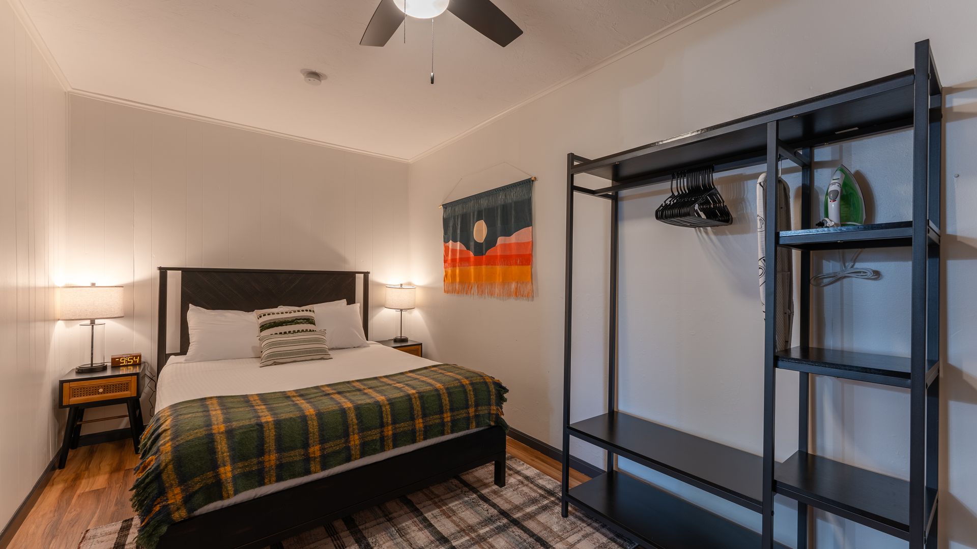 A bedroom featuring a bed with a plaid blanket, a wooden nightstand with a lamp, and an open metal clothing rack.