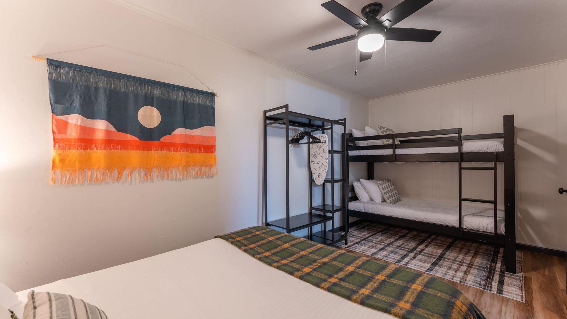 A bedroom featuring a bunk bed, a metal clothing rack, and a wall hanging with sunset colors above a bed with green bedding.