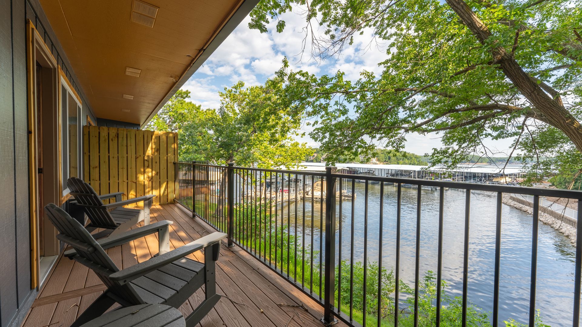 A balcony with two dark chairs overlooking a lake and marina, framed by lush trees under a partly cloudy sky.