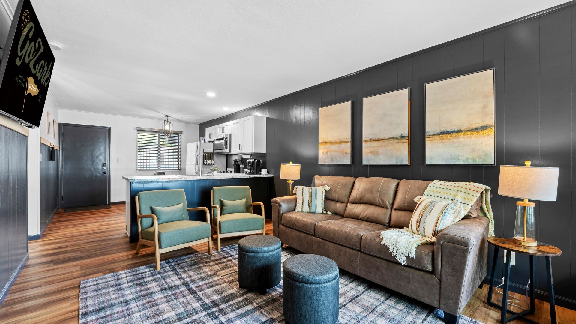 A modern living room with a brown sofa, two sage green chairs, a dark accent wall, and an open kitchen in the background.