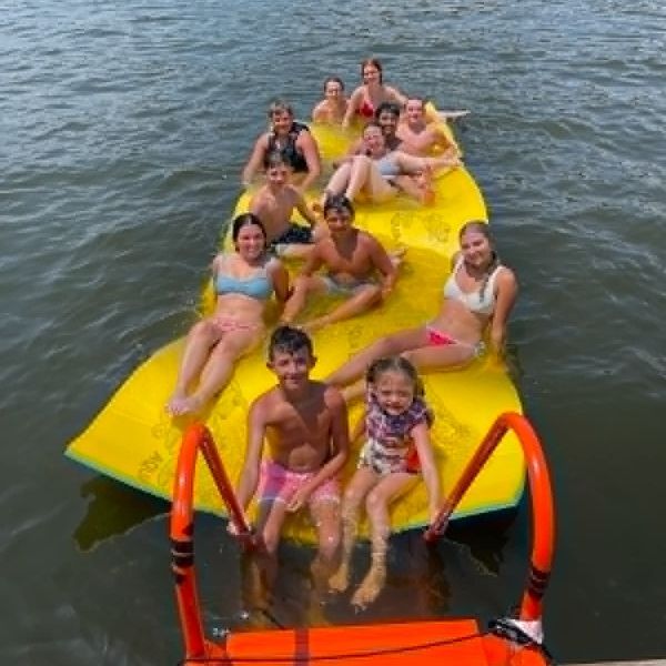 People lounging on a bright yellow water trampoline in a lake, smiling and splashing.