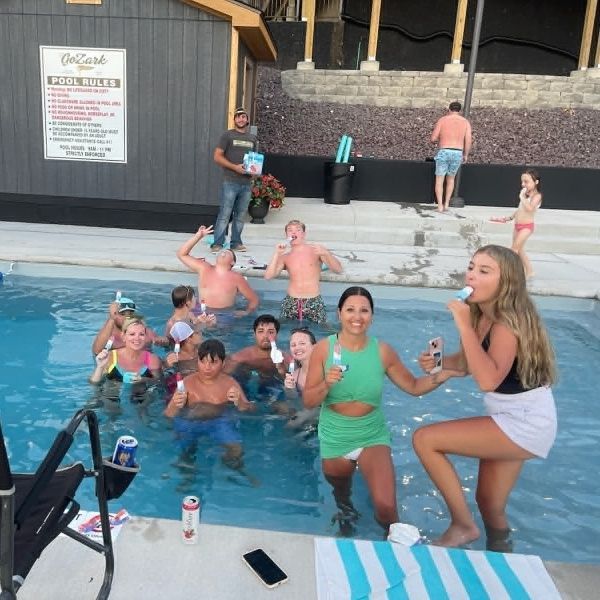 People posing and playing in a pool at a lively outdoor gathering, with a stage and building in the background