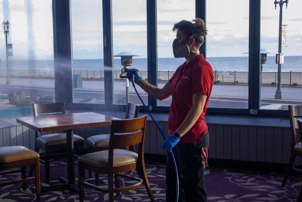 A person in red shirt and mask sprays disinfectant inside a restaurant near windows with a beach view.