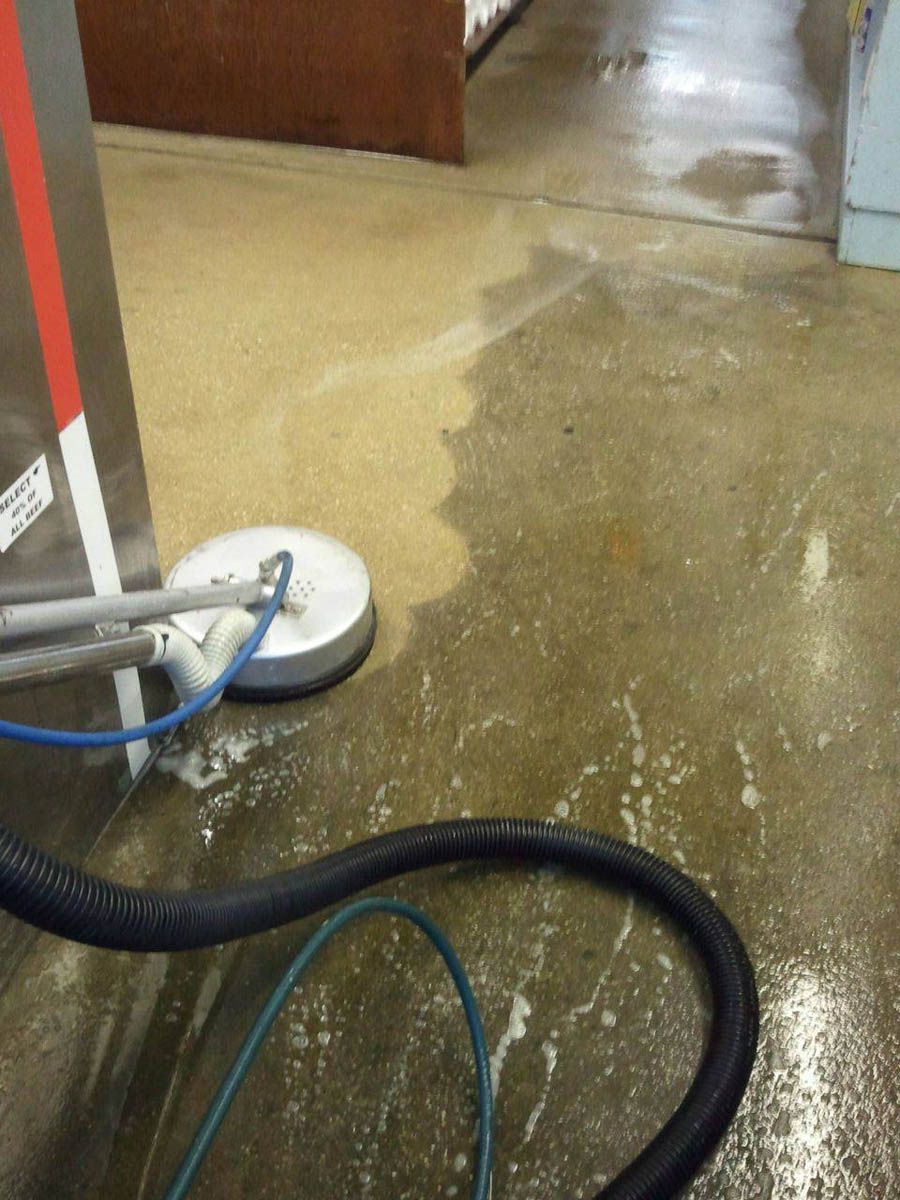 A floor cleaning machine in use, revealing a clean strip on a concrete floor; soapy water and a black hose visible.